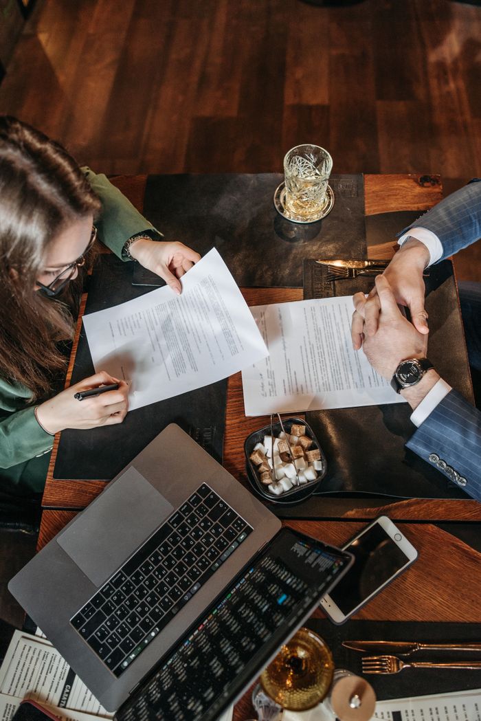 О нас High angle shot of a business meeting with a woman signing a contract on a table with a laptop.