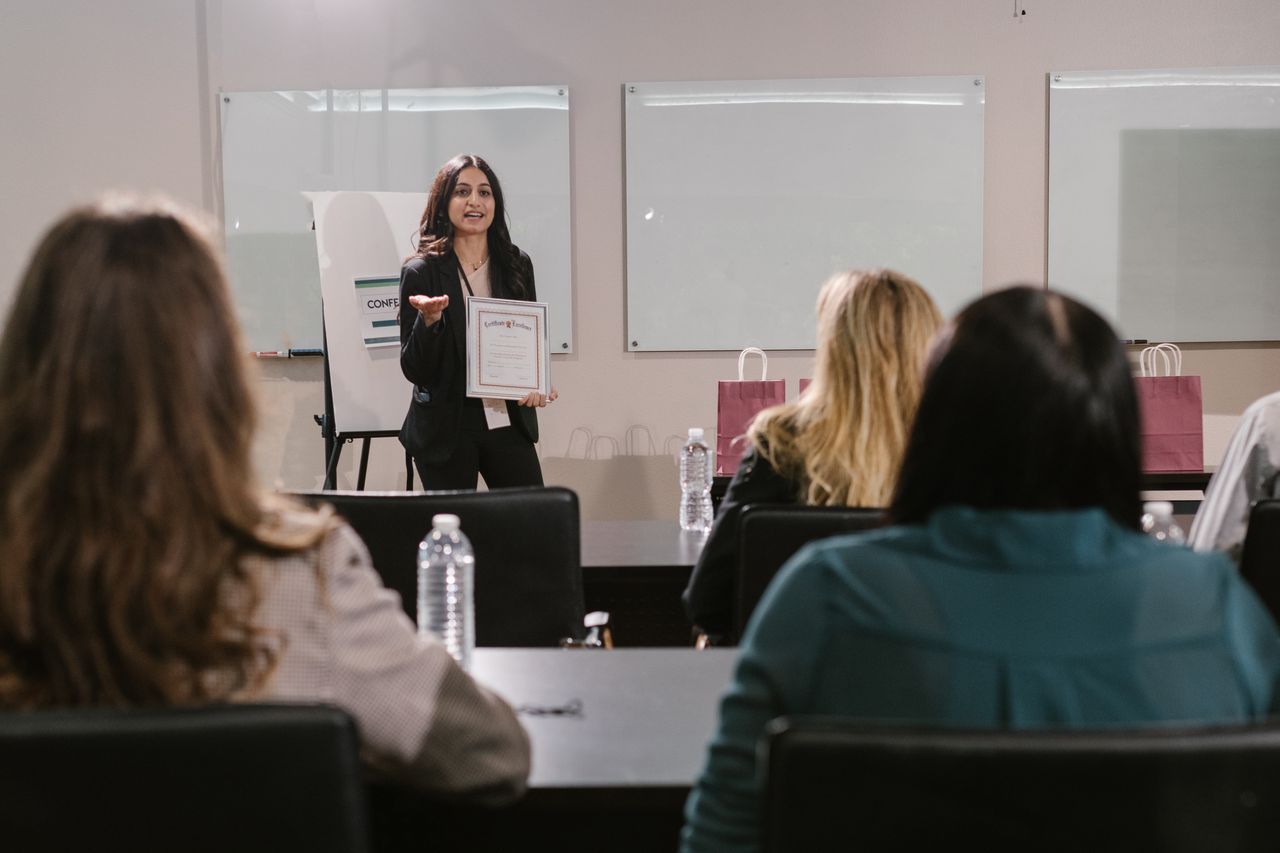 Home A business seminar showcasing a female presenter awarding certificates to attendees.