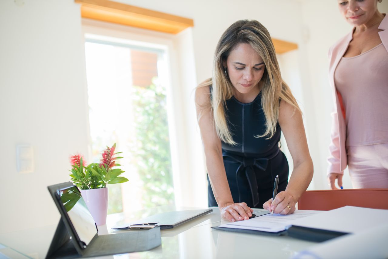 Home Businesswomen signing a contract at a bright office desk with documents and tablet.